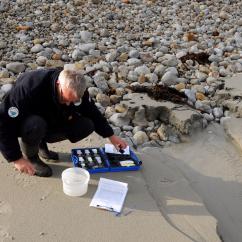 Un agent accroupi sur une plage analyse les pollutions d'un filet d'eau se déversant dans la mer